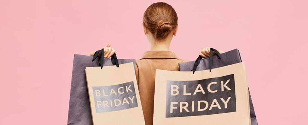 Mulher de blazer e cabelo apanhado, de costas, segura sacos de compras a dizer "Back Friday". Fundo rosa